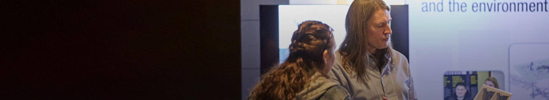 Photograph: two people talk at an exhibit; one holds a brochure and gestures by a poster.